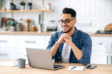 Photo of attractive positive indian or arabian guy with glasses, student or freelancer, working remotely, sitting at home in the kitchen with laptop, looking at the screen, thinking, smile happily