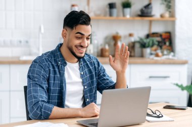 Online video communication. Friendly positive freelancer or student, arabian or indian nationality, sits at a desk at home, talking on video call with colleagues, greeting with hand gesture, smiling