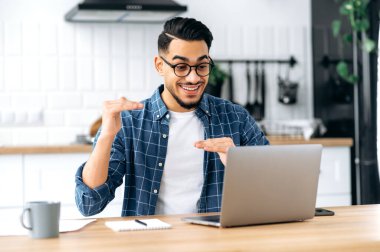 Distant communication. Friendly arabian or indian man with glasses, sitting at home in the kitchen, holding video call with client, virtual meeting with colleague, uses laptop, gesturing hands, smile