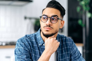 Close-up photo of a serious modern Indian or Arabian young man with glasses, with a beard, stylishly dressed, student, freelancer, IT specialist, looking away, thinking, planning, dreaming