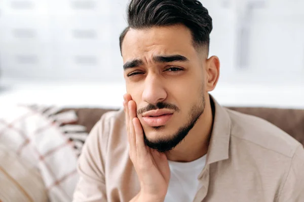 Frustrated unhappy indian or arabian man in casual clothes, pressing cheek with hand, suffering toothache, caries, sitting of sofa in living room, needs the help of a dentist. Dental problems concept