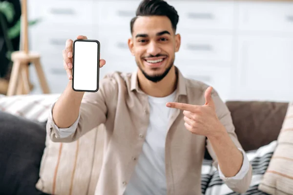 Phone screen mockup. Defocused smiling mixed race guy, showing smart phone with empty white mock-up screen, copy-space for advertising or presentation, sitting on a sofa in a living room interior
