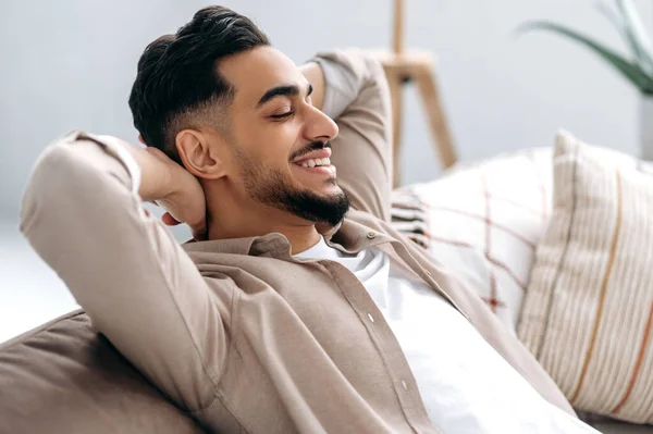Chilling and relaxing at home. Handsome happy calm indian or arabian guy in casual clothes resting in the living room on the sofa, holds his hands behind his head with closed eyes and smiles