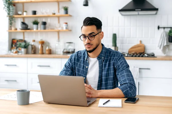 Serious focused arabian or indian guy with glasses, in stylish casual clothes, student, freelancer, company employee working remotely, sitting in the kitchen with a laptop, working on a project