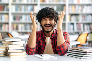 Puzzled confused indian or arabian guy, a university student, sit at a desk in library between books, looking at the camera in disappointment spreading his arms to the sides, emotional face expression