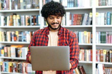 Stylish modern curly haired positive arabian or indian university student, holding an open laptop in his hands, standing in the library, browsing the internet, looking for information, smiling