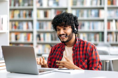 Friendly stylish Indian or Arabian guy, male student or freelancer with headset, sitting at a desk in a library, using laptop for online conversation, discuss information with interlocutor, smiling