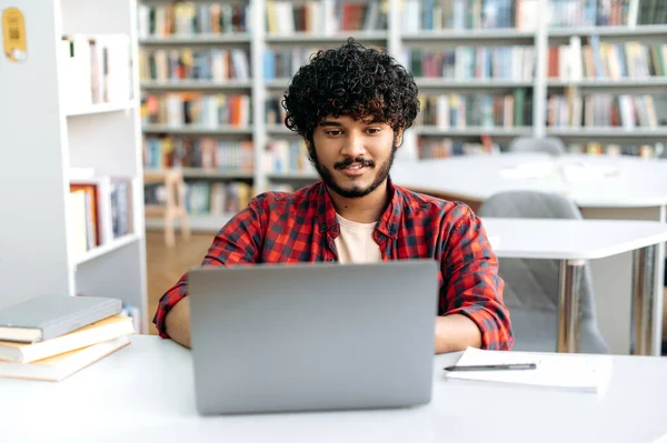 Clever confident positive arabian or indian male student, stylishly dressed, sit at table with laptop in university library, working on a project, prepares homework, browsing internet resources, smile