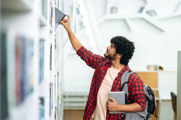 Choosing a book. Clever arabian or indian male student, in casual stylish wear, with a backpack, stands in the library near the bookshelf, chooses a book for himself, takes it from the shelf, smiles