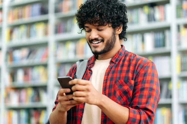 Happy indian or arabian millennial guy, freelancer or student, using his smartphone, chatting with friends on social networks, texting sms, browses internet,smile friendly. Wireless technology concept