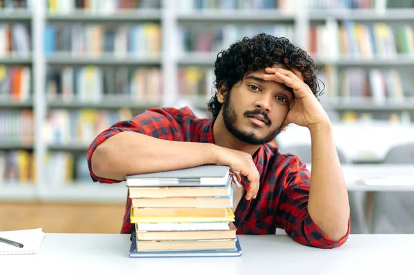Tired exhausted curly haired indian or arabian guy, mixed race male, university student, sits at a desk with books, in the library against the background of bookshelves, looks at camera