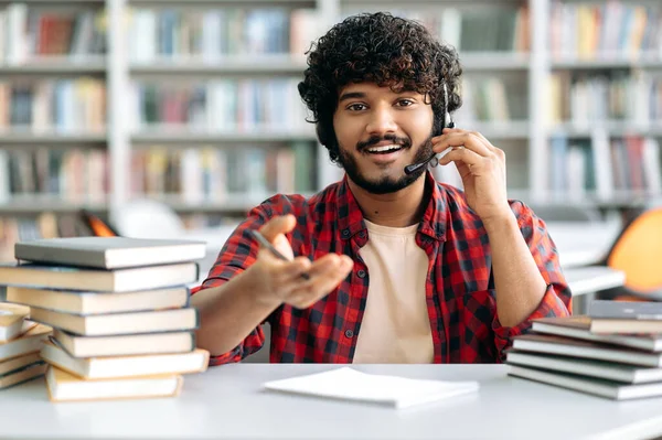 Positive Arabic or Indian university student with curly hair, wearing a headset, sits at a table in the library with books, watching an online webinar, listening to a lecture, looks at camera, smiling