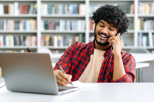 Phone talk conversation during work or study. Positive busy indian or arabian curly haired happy guy, having negotiation by smart phone, talks with friend or colleague, sits at desk, smiling friendly