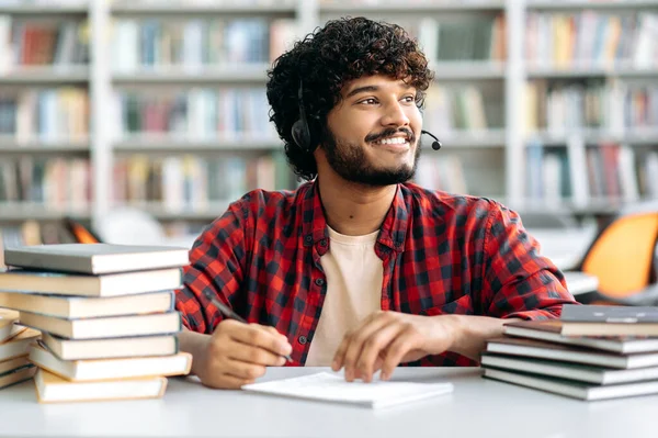Positive Arabian or Indian university student with curly hair, with a headset, sits at a table in the library with books, listening an online webinar, lecture, looks away, dreaming, thinking, smiling