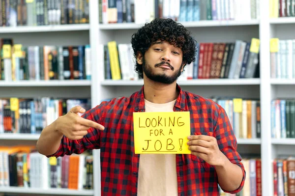 Upset curly indian or arabian unemployed guy, stands in the library against the background of bookshelves, holds a sign in his hand that says looking for a job, looks at camera, hopes to get a job