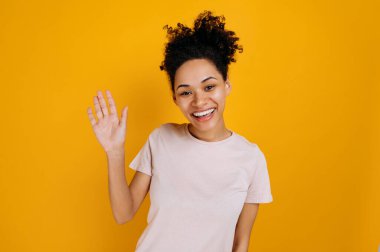 Greeting gesture. Positive pretty, curly haired african american young woman, waving hand, makes greeting gesture, looking at the camera, smiling, standing over isolated orange background