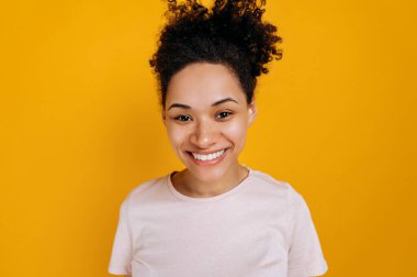 Close-up photo of happy african american young woman with curly hair and freckles, wearing basic t-shirt, standing on isolated orange background, looking straight at camera, smiling friendly