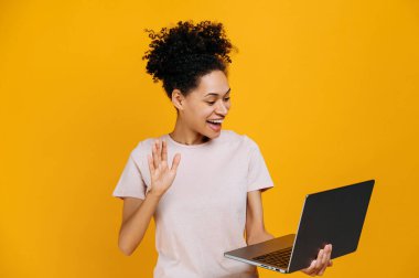 Online video communication. Friendly african american woman, holding open laptop in hand, doing hello gesture, talking on video conference, standing over isolated orange background, smiling