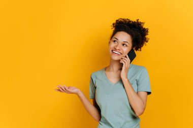 Phone talk by mobile phone. Happy excited african american young woman has pleasant phone conversation, happy facial expression, stands on isolated orange background, looks away, gesturing hand, smile