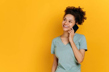 Pleasant phone conversation. Positive african american curly young woman in basic t-shirt, is talking on the smartphone with her friend or family, smiling, standing on isolated orange background