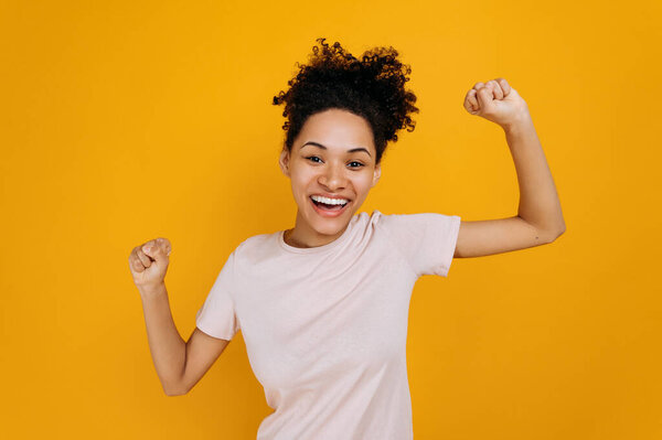 Радостная счастливая кудрявая африканская американка, танцующая, жестикулирующая кулаками, получает прибыль, рада выиграть в лотерею, стоя на изолированном оранжевом фоне. Хорошие новости, успех и счастье