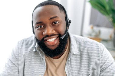 Close-up of a friendly positive african american man with headset, call center worker, expert, company representative, support service operator, holds online consultation, looks at camera, smiles