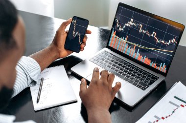 Crypto trader investor broker using cellphone and laptop for cryptocurrency financial market analysis, buying or selling cryptocurrency. Close-up of a laptop and cellphone screen with stock diagrams