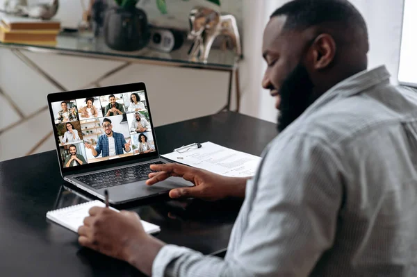 Group brainstorm, online video meeting, virtual conference with multiracial colleagues. African american man talking with colleagues by video call uses laptop and app, takes notes, discuss ideas