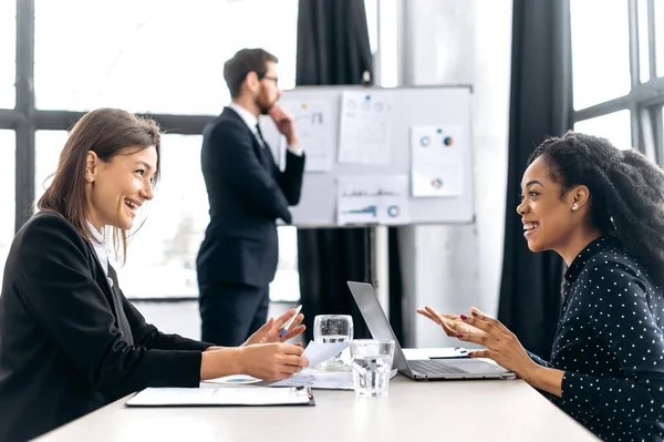 Two successful confident women are negotiating, discussing business project, planning financial strategy, smile. Elegant man stands on the background near white board, thinking business plan