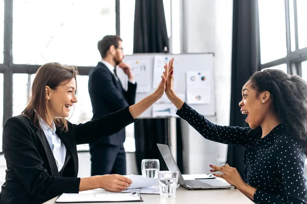 Two successful multi-ethnic female colleagues work in a modern office on a project, give each other five, rejoice at a successful deal, good profit, a successful startup, smiling. Agreement, deal