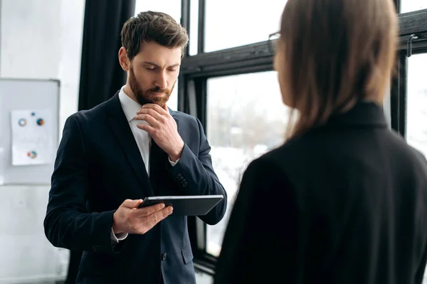 Two successful company colleagues, caucasian man in a formal suit, communicates with his female colleague, they stand in the office, discuss work issues, financial reports, company profits