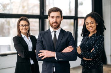 Successful business partners. Multiracial successful colleagues, dressed in formal wear, standing in their modern office with their arms crossed, looking at the camera, smiling friendly