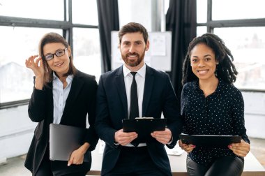 Elegant creative colleagues stand in modern office, show confidence and success, teamwork, collaboration concept. Multiracial influential business people in formal wear look at the camera, smiling