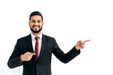 Friendly positive confident indian or arabian businessman in a black business suit, standing over isolated white background, pointing fingers to the side, looking at camera, smiling happily