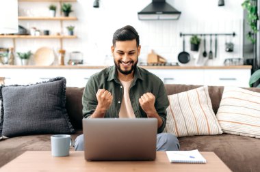 Win, success, triumph. Happy arabian and indian man, sits on a sofa in living room, looks at laptop screen, gesticulates with fists, celebrates success, victory, big profit, received good news, smiles