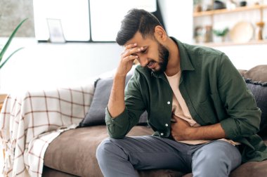 Unhappy indian or arabian man, sits on comfortable sofa in cozy living room, holds his hands on his stomach, grimaces from pain in his stomach, suffers from poisoning, spasm, stomach problems