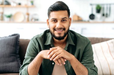 Portrait of confident attractive indian or arabian man, in casual stylish clothes, sitting at home in cozy living room on the background of the kitchen, looks at camera, smiling friendly