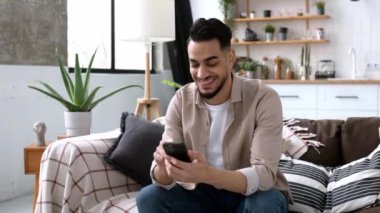 Çekici, dost canlısı Hintli ya da Arap bir adam, şık giyinmiş, oturma odasındaki koltukta oturuyor, akıllı telefonunu kullanıyor, arkadaşlarıyla sosyal medyada sohbet ediyor, online alışveriş yapıyor.