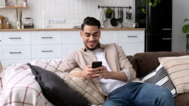 Attractive indian or arabian young man, sit in a modern living room on comfortable sofa, uses his smartphone, texting message, browsing social media, looks away, smiles. The concept of using gadgets