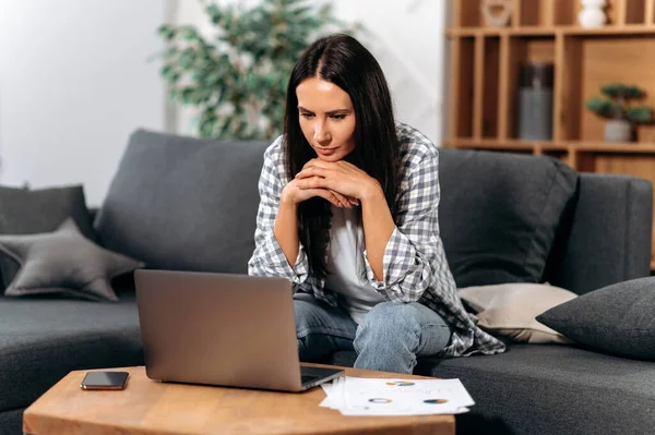 Focused confident caucasian brunette woman, sit in living room, using laptop, working on an important project from home, concentrated looks at screen, looking for information on the internet