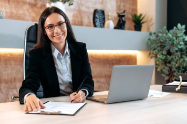 Portrait of confident Caucasian female broker in the office. Beautiful successful smart brunette woman, top company manager or trader, uses laptop, study documents, looks at camera, smiling