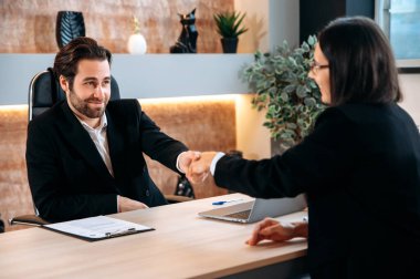 Cooperation, agreement. Successful caucasian business colleagues, entrepreneurs, directors, sit at a desktop in modern office, made a good deal, agreed of cooperation, shake hands to each other, smile
