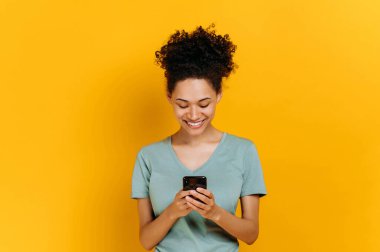 Wireless technology, gadget. Excited positive african american curly young woman uses smartphone, browsing internet, messaging with friends online, stand on isolated orange background, looks at screen