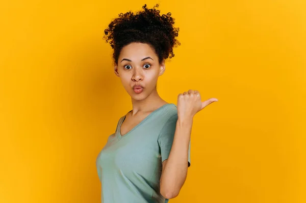 Surprised emotion african american curly haired young woman in a t-shirt, points finger to the side behind the back, at empty copy space, stand on isolated orange background, shocked looks at camera
