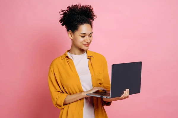 Positive lovely african american modern young curly haired woman, holding an open laptop in hand, looks at the screen, standing against isolated pink background, smiling happily. Wireless technology