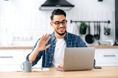 Video communication. Friendly arabian or indian man with glasses, sitting at home in the kitchen, conducts video call with client, virtual meeting with colleague, uses laptop, greeting gesture, smiles