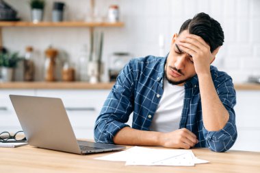 Tired worried sad arabian or indian guy, sit at a desk, looking tired and stressed, frustrated looking at work documents, failing to finish work on the project on time, needs rest. Overworking concept