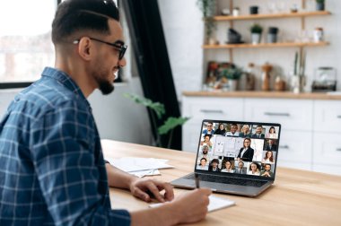 Online education. View over the shoulder of a mixed race guy taking notes in a notebook, at a laptop screen with a teacher and a group of multiracial students. E-learning, webinar, online lecture