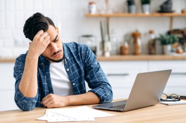 Sad arabian or indian guy, sitting at a desk, looking tired and stressed, frustrated looking at work graphs, failing to finish work on the project on time, needs rest. Overworked from work, deadline