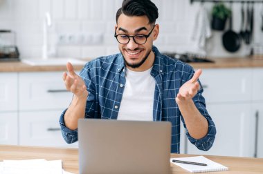 Amazed Arabian or Indian man with glasses, mixed race clever student or freelancer, having distant communication by video call uses laptop, listening to an online webinar, lecture, smiling friendly
